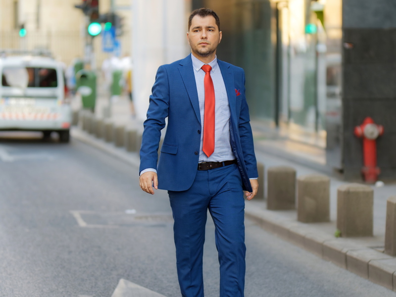 A man wearing a royal blue textured suit with a patterned shirt and a bright red tie while walking down an urban street